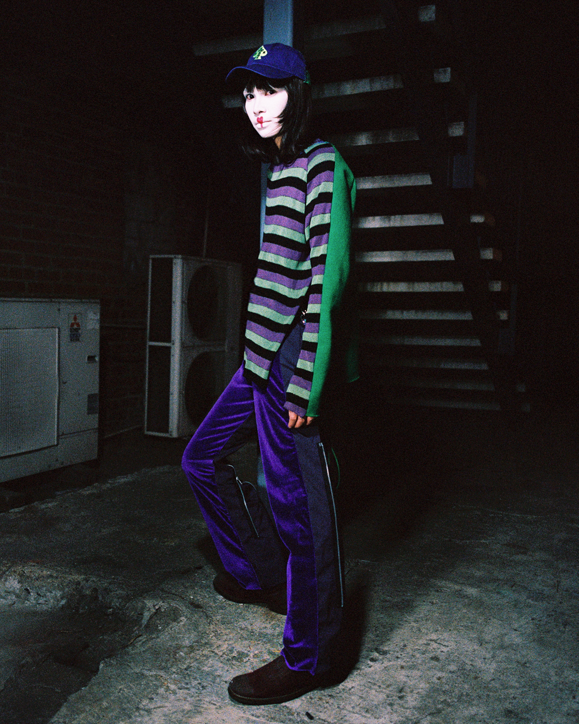 Model with white facepaint with graphic red details standing contraposto under a staircase